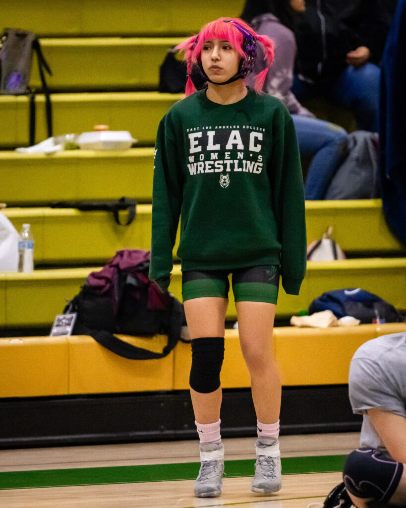 Image of Mollie Jimenez, 109-pound weight class wrestler warming up before her bout with Amber Chatterton. Jimenez wears a green, oversized "ELAC Women's Wrestling" sweatshirt and jumps up and down. 