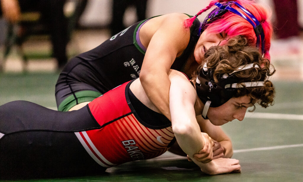 A wrestler is face down on the mat, but with her arms pushing her upper body off the mat as another wrestler holds her from above. 
